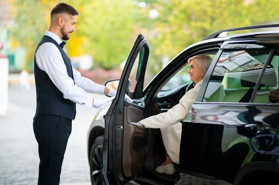 A man dressed in a suit is opening a black car door, symbolizing a chauffeur service near me.